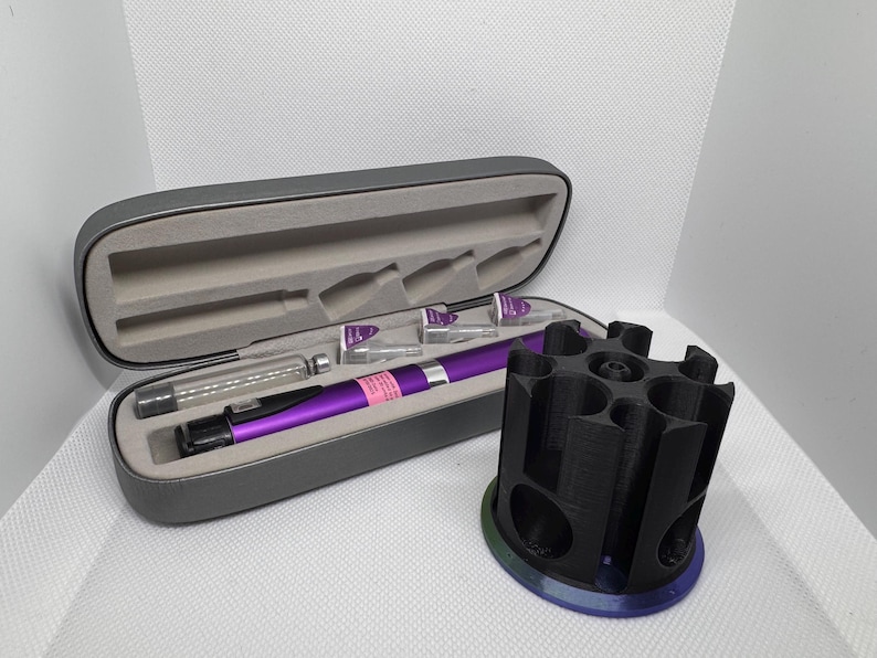 Puede incluir: Un bol&iacute;grafo m&eacute;dico morado y accesorios se muestran en un estuche gris abierto. A la derecha hay un soporte cil&iacute;ndrico negro con m&uacute;ltiples aberturas. El estuche contiene un bol&iacute;grafo, viales y otros suministros m&eacute;dicos.