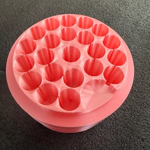May include: A coral-colored, circular organizer with multiple cylindrical holes. The organizer has a tiered design, with a slightly raised base and a top section filled with evenly spaced openings. The object is made of a solid material.