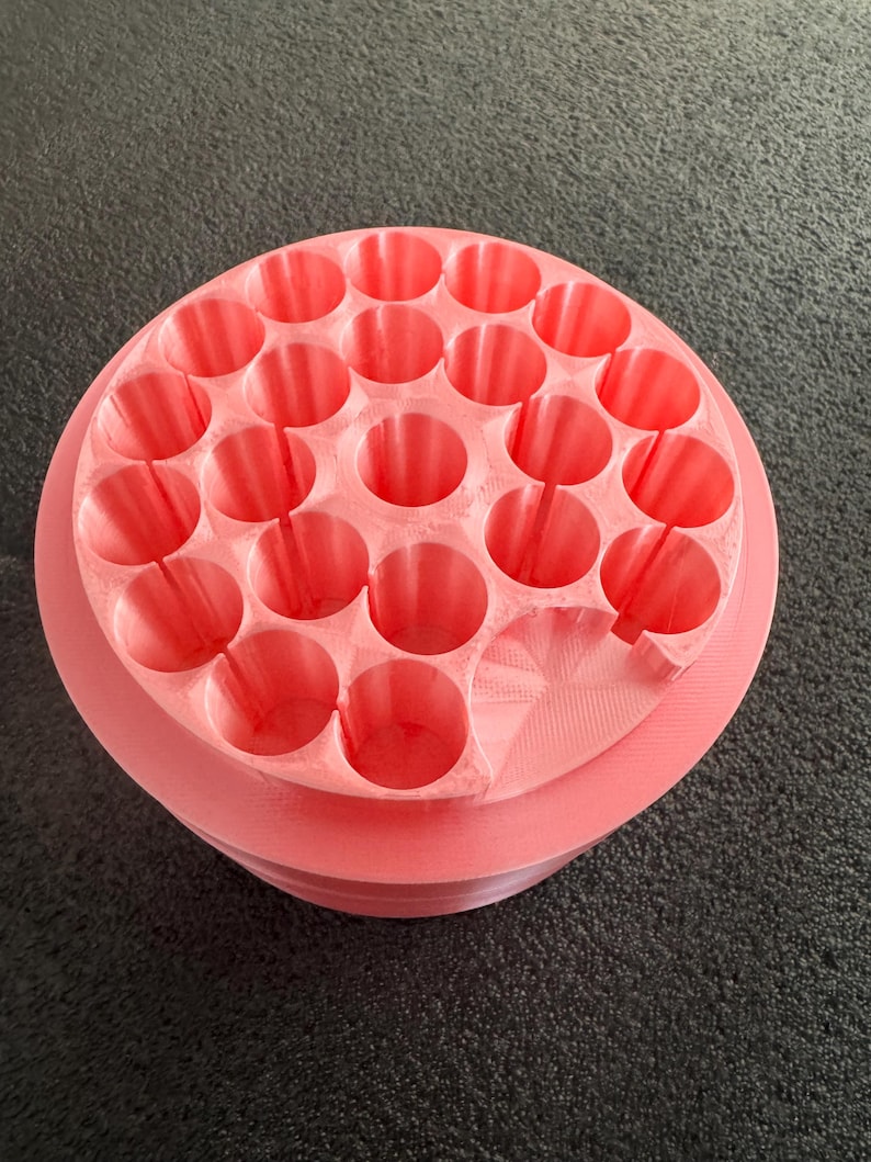 May include: A coral-colored, circular organizer with multiple cylindrical holes. The organizer has a tiered design, with a slightly raised base and a top section filled with evenly spaced openings. The object is made of a solid material.