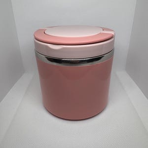 May include: A pink and white insulated food container. The container has a cylindrical shape with a stainless steel band. The lid is light pink with a darker pink accent. The container is designed to keep food warm or cold.