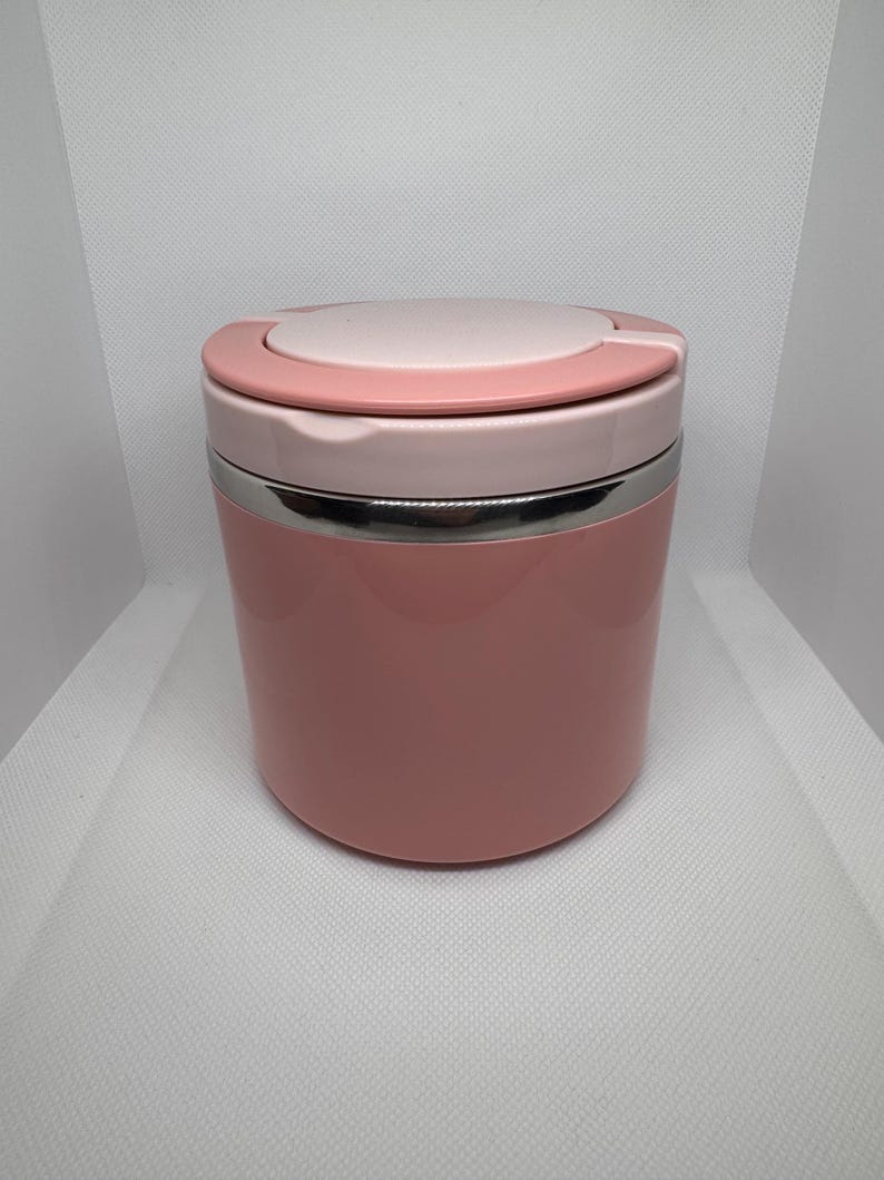 May include: A pink and white insulated food container. The container has a cylindrical shape with a stainless steel band. The lid is light pink with a darker pink accent. The container is designed to keep food warm or cold.