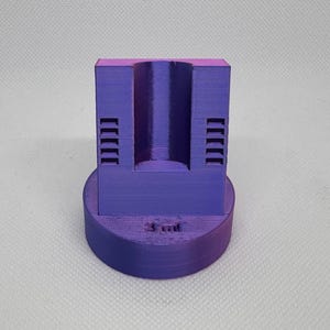 May include: A 3D-printed purple object with a cylindrical hole and a circular base. The object has a rectangular top section with a series of parallel lines on each side. The base has the text "3 ml" printed on it.