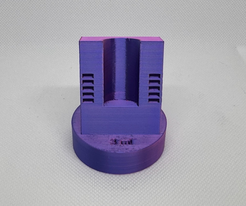 May include: A 3D-printed purple object with a cylindrical hole and a circular base. The object has a rectangular top section with a series of parallel lines on each side. The base has the text "3 ml" printed on it.