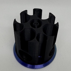 Puede incluir: Un objeto cil&iacute;ndrico negro impreso en 3D con m&uacute;ltiples aberturas circulares y una base azul. El objeto tiene un dise&ntilde;o geom&eacute;trico complejo, con una serie de bordes curvos y un agujero central. La base es de forma circular s&oacute;lida.