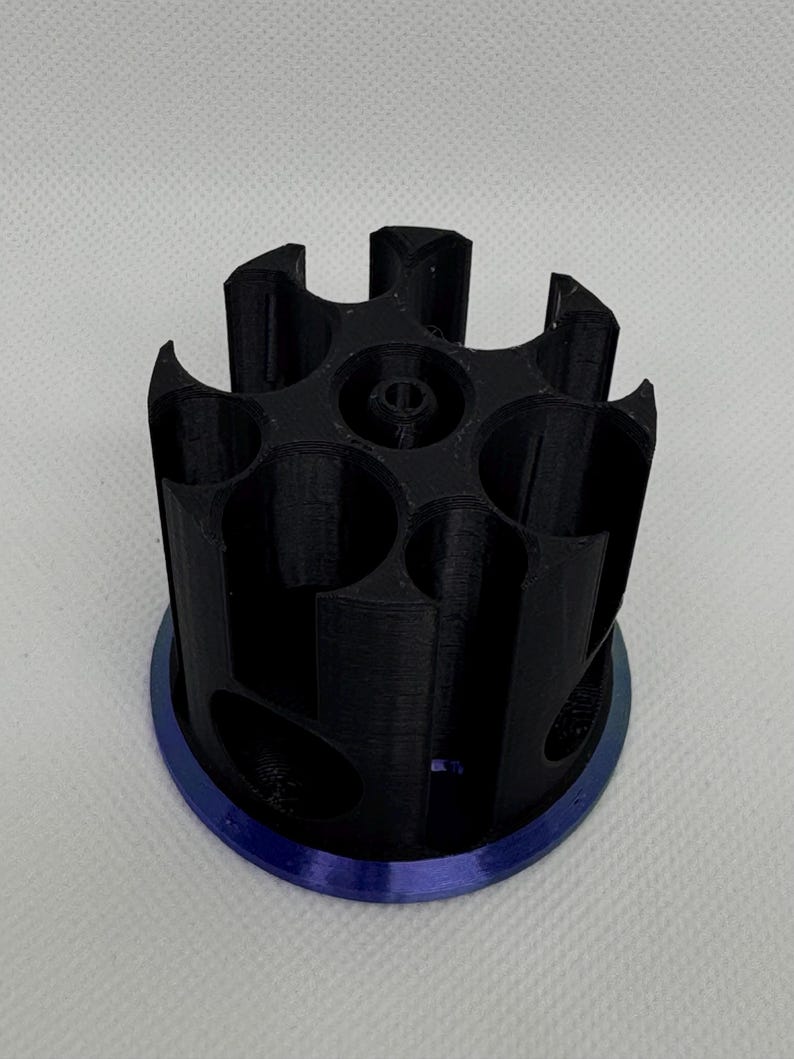 Puede incluir: Un objeto cil&iacute;ndrico negro impreso en 3D con m&uacute;ltiples aberturas circulares y una base azul. El objeto tiene un dise&ntilde;o geom&eacute;trico complejo, con una serie de bordes curvos y un agujero central. La base es de forma circular s&oacute;lida.