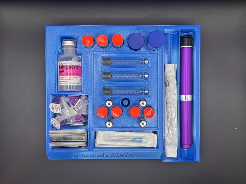 Puede incluir: Una bandeja azul con varios suministros m&eacute;dicos. Incluye una botella etiquetada como "Agua Bacteriost&aacute;tica", jeringas, agujas y viales con tapas rojas y azules. Tambi&eacute;n hay un dispositivo morado en forma de bol&iacute;grafo.
