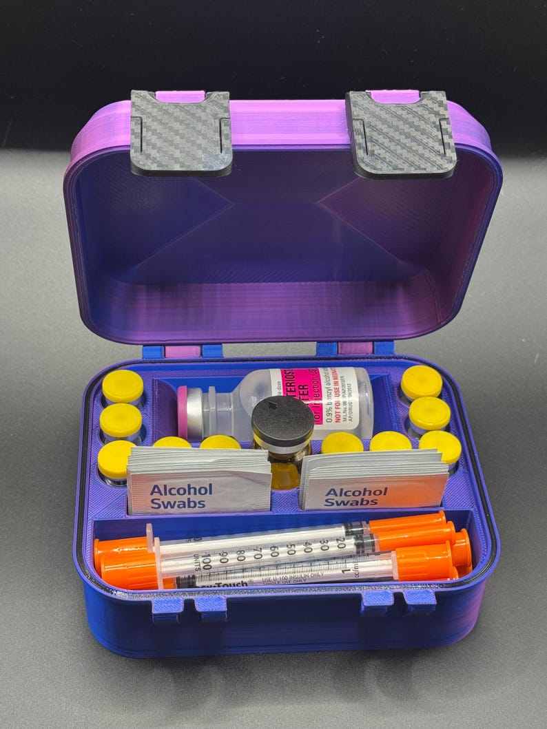 May include: A purple storage case with two carbon-fiber patterned latches containing medical supplies. The interior features organized compartments holding a glass vial, several small yellow-capped bottles, two stacks of alcohol swabs, and syringes.