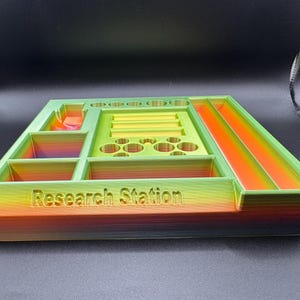 Puede incluir: Un organizador de Research Station multicolor impreso en 3D. El organizador tiene varios compartimentos, incluyendo ranuras, bandejas y soportes circulares. Los colores transicionan de verde a amarillo a naranja, con el texto "Research Station".