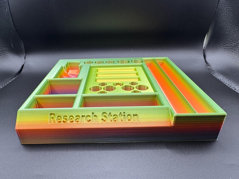 Puede incluir: Un organizador de Research Station multicolor impreso en 3D. El organizador tiene varios compartimentos, incluyendo ranuras, bandejas y soportes circulares. Los colores transicionan de verde a amarillo a naranja, con el texto "Research Station".