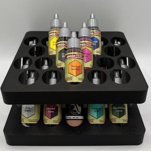 Könnte beinhalten: Ein schwarzer, zweistöckiger Farbflaschen-Organizer, gefüllt mit verschiedenen Speed Paint-Flaschen. Die Flaschen sind mit Farben wie Nuclear Sunrise, Holy White und Absolution Green beschriftet. Der Organizer ist für mehrere Farbflaschen konzipiert.