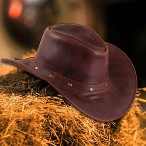 May include: A brown leather cowboy hat with a wide brim and a rounded crown. The hat features a decorative band with small, evenly spaced rivets. The hat is resting on a bed of hay, suggesting a rustic or Western theme.