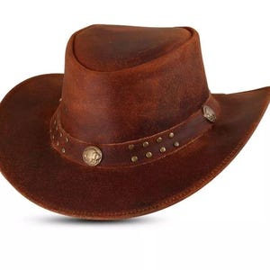 May include: A brown leather cowboy hat with a wide brim and a rounded crown. The hat features a decorative band with small, round studs and two circular metal embellishments. The hat is designed in a classic Western style.