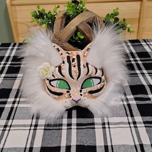 May include: A cat mask featuring a tan base with black stripes and white spots, accented by long white faux fur trim around the edges. The mask includes bright green mesh eye coverings, a small pink nose, and a single cream-colored fabric flower.