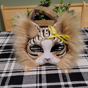 May include: A cat mask featuring a white base with brown and black tabby stripes, accented by long tan faux fur trim around the edges. The mask includes black eye cutouts, a small pink nose, and a bright yellow ribbon bow attached to one ear.