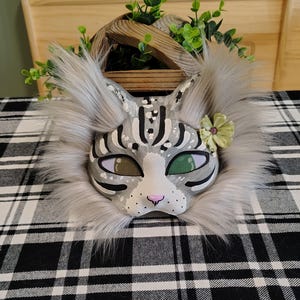 Op de afbeelding: Een kattenmasker met grijs en wit bont, geschilderde grijze en witte strepen, een kleine roze neus en groene mesh oogbedekkingen. Een enkele lichtgroene stoffen bloem is aan de zijkant van het masker bij het oorgedeelte bevestigd.