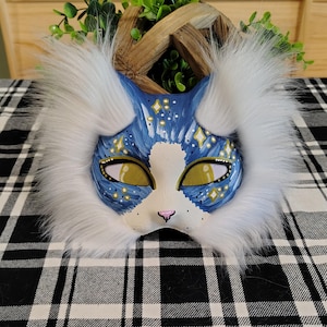 Op de afbeelding: Een katvormig masker met een blauw geschilderd gezicht met witte accenten, gouden sterdetails en gele mesh oogbedekkingen. Het masker is omlijst door dik, lang wit imitatiebont aan de zijkanten en bovenkant, met een kleine roze neus in het midden.