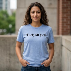 Puede incluir: Una camiseta azul claro con la frase "Fuck All Ya'll" en negro. La camiseta de manga corta parece estar hecha de un material suave. La persona lleva la camiseta y tiene las manos en los bolsillos.