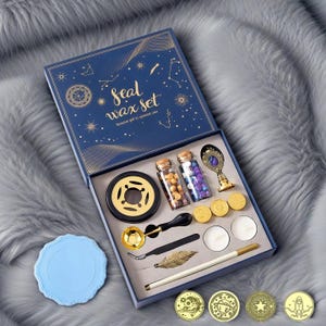 Puede incluir: Un juego de sellado de cera azul marino con detalles dorados. La caja abierta revela cuentas de cera, un crisol, una cuchara, un bolígrafo y sellos de cera. La tapa tiene las palabras "Seal Wax Set" y diseños de constelaciones.