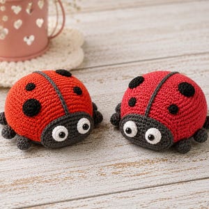 May include: Two handmade crochet ladybirds featuring bright red bodies with black spots, dark grey heads, and white eyes with black pupils. Each ladybird has small dark grey crocheted legs extending from the sides of their rounded, textured bodies.