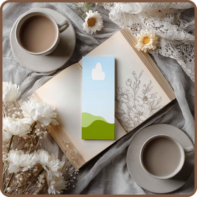 May include: A bookmark with a landscape design featuring a blue sky, white cloud, and green hills, resting on an open book with floral illustrations. Two cups of coffee and white flowers are arranged around the book.