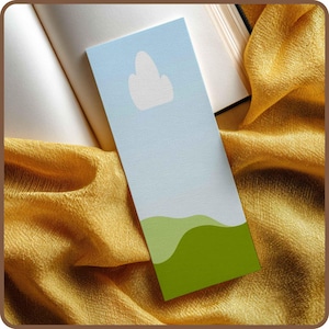May include: A rectangular bookmark with a light blue sky design and a white cloud. The bottom of the bookmark features green rolling hills. The bookmark is resting on an open book and a gold fabric.