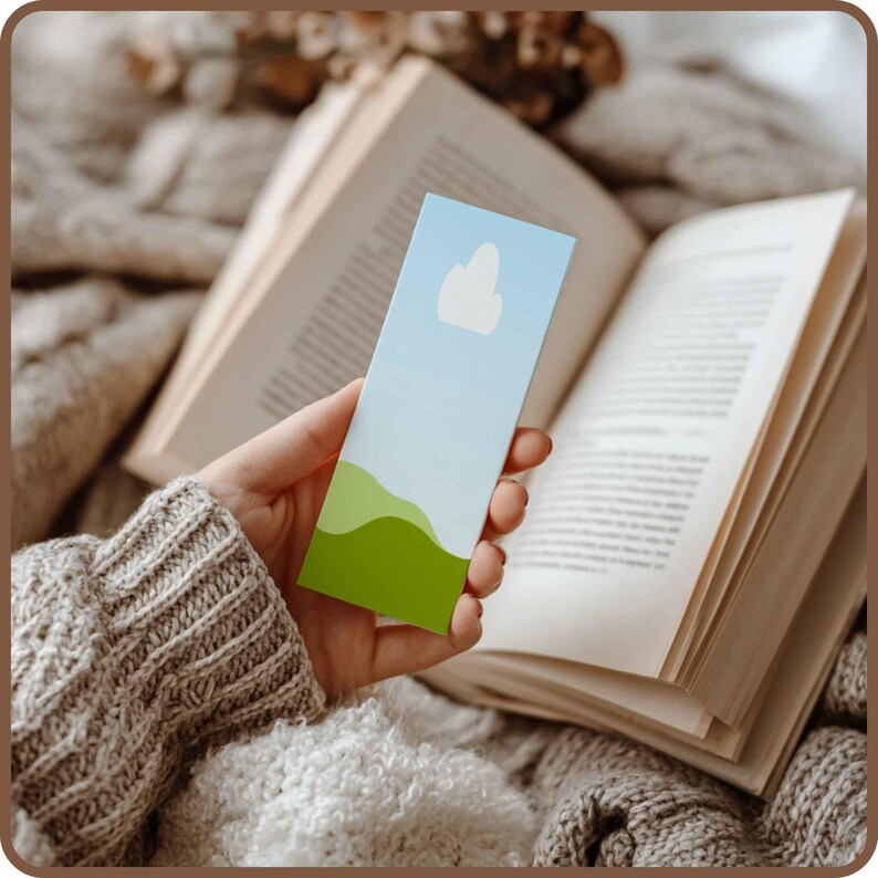 May include: A rectangular bookmark with a sky-themed design is held in front of an open book. The bookmark features a gradient blue sky, a white cloud, and green rolling hills. The book has aged, off-white pages.