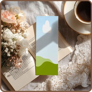 May include: A colorful bookmark with a sky and landscape design. The bookmark features a light blue sky with a white cloud and green rolling hills. The bookmark is placed on an open book, next to a bouquet of flowers and a cup of coffee.