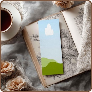May include: A bookmark with a minimalist design featuring a blue sky, a white cloud, and green rolling hills. The bookmark is set against an open antique book with floral illustrations, a cup of tea, and flowers.