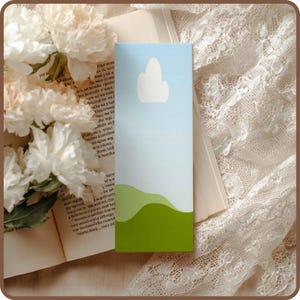 May include: A rectangular bookmark with a landscape design. The top portion is light blue with a white cloud shape. The bottom has green rolling hills. The bookmark is resting on an open book with white flowers.