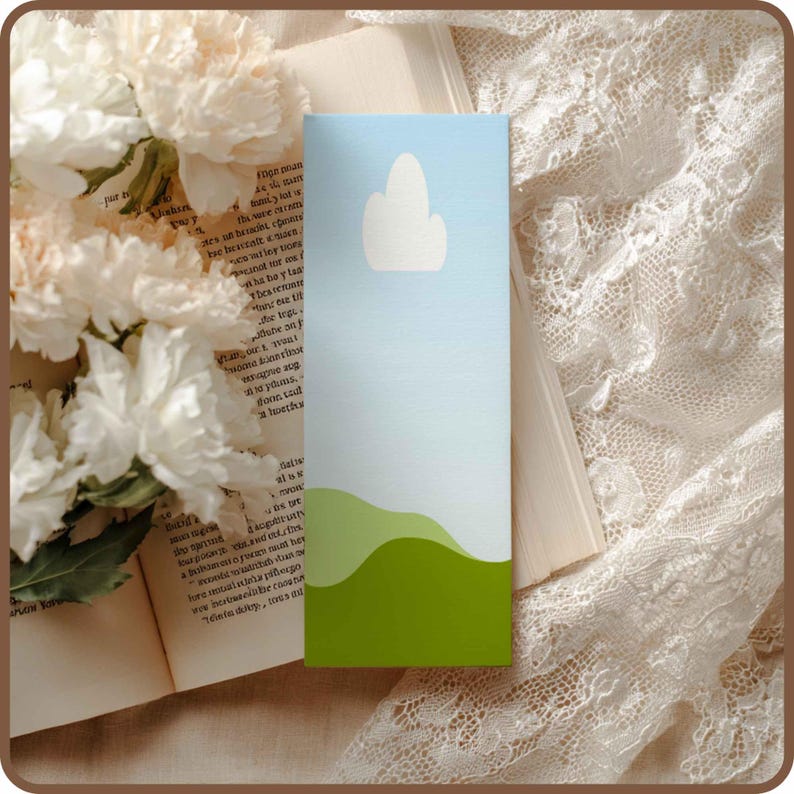 May include: A rectangular bookmark with a landscape design. The top portion is light blue with a white cloud shape. The bottom has green rolling hills. The bookmark is resting on an open book with white flowers.
