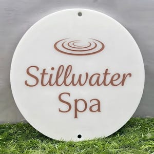 Pode incluir: Uma placa circular branca com o texto Stillwater Spa numa fonte serifada castanha. Acima do texto, existe um gráfico castanho de círculos concêntricos que representam ondulações. A placa tem um orifício no topo e na base.