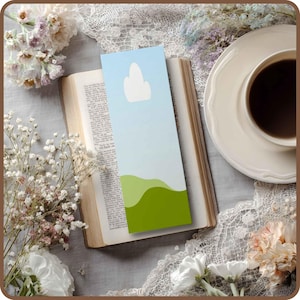 May include: A colorful bookmark with a sky and landscape design is placed in an open book. The bookmark features a light blue sky with a white cloud and green rolling hills. A cup of coffee sits on a white saucer.