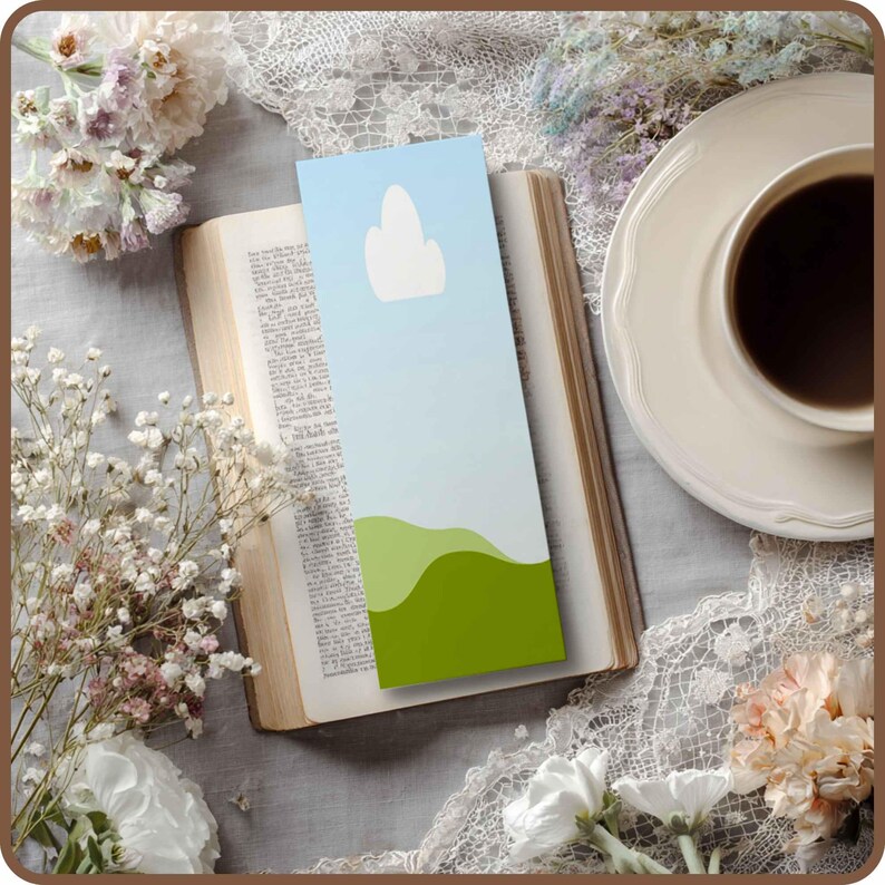 May include: A colorful bookmark with a sky and landscape design is placed in an open book. The bookmark features a light blue sky with a white cloud and green rolling hills. A cup of coffee sits on a white saucer.