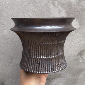 Peut inclure: Un vase en céramique brun foncé avec un motif texturé et nervuré. Le vase a un bord large et évasé et un corps cylindrique. L'extérieur a un aspect rugueux et fait à la main, adapté à la décoration intérieure.