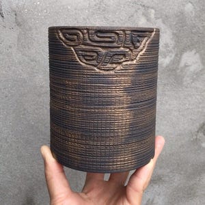 May include: A cylindrical, dark brown ceramic pot with a textured, grid-like pattern. The top edge features an embossed, stylized design. The pot is held in a hand against a gray background, showcasing its unique texture and form.