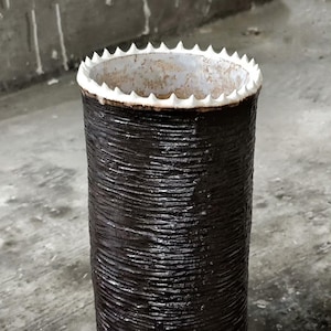 Op de afbeelding: Een cilindervormige, donkerbruine vaas met een getextureerd, geribbeld oppervlak. De bovenrand is wit met een gekarteld, tandachtig ontwerp. De vaas is een decoratief item, waarschijnlijk voor de inrichting.