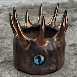 May include: A handcrafted, cylindrical ceramic container with a crown-like design. The container is dark brown with bronze-colored spikes. A blue and white eye-shaped embellishment is on the side.