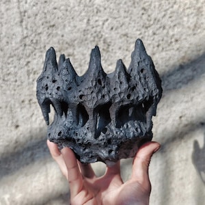 Puede incluir: Una maceta decorativa de color gris oscuro y texturizada con un diseño único, dentado y poroso. La maceta tiene un aspecto rugoso y orgánico, con bordes irregulares y una serie de pequeños agujeros en su superficie. La maceta se sostiene en una mano.