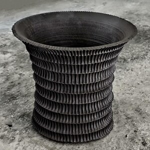 Könnte beinhalten: Eine dunkelgraue, strukturierte Keramikvase mit breitem, geriffeltem Rand und zylindrischem Körper. Der Körper weist eine Reihe vertikaler, geriffelter Muster auf, die einen geschichteten Effekt erzeugen. Die Vase ist etwa 25 cm hoch.