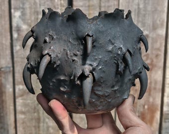 May include: A dark gray, textured ceramic bowl with a rounded shape. The bowl's rim features jagged edges and is adorned with several claw-like protrusions. The bowl is held in a hand against a wooden background.