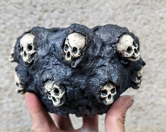 May include: A dark gray, textured, skull-adorned planter. The planter features multiple off-white skulls embedded in the dark gray exterior. The skulls are evenly spaced around the planter's rim, creating a gothic or Halloween theme.