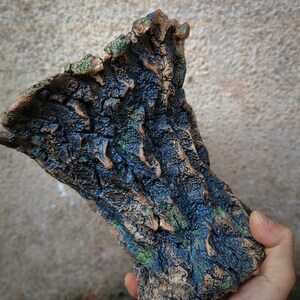 May include: A textured, hand-held decorative piece resembling a section of tree bark. The object features a mix of dark blue, green, and brown hues, with a rough, uneven surface. The bark-like design is detailed and naturalistic.
