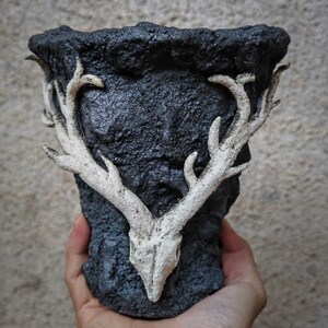 May include: A black, textured ceramic planter with a white deer skull and antler design. The planter has a rough, stone-like appearance and is held by a hand. The antlers are prominently displayed on the front of the planter.