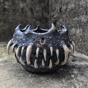 Pode incluir: Uma tigela de cerâmica escura e texturizada com uma borda irregular e uma fileira de dentes grandes e esbranquiçados na parte externa. A tigela tem um pequeno detalhe retangular na lateral, evocando a boca de uma criatura.