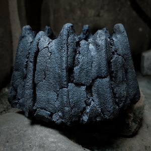 Könnte beinhalten: Ein dunkelgrauer, strukturierter Keramik-Pflanztopf mit einem gezackten, bergähnlichen Rand. Der Pflanztopf steht auf einer Steinoberfläche und zeigt sein einzigartiges, organisches Design. Die Außenseite des Pflanztopfs hat ein raues, verwittertes Aussehen.