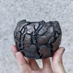 Può includere: Un piccolo vaso rotondo in ceramica scura con una superficie strutturata e screpolata. Il vaso ha una lucentezza metallica ed è tenuto in mano su uno sfondo neutro. Il design del vaso suggerisce un'estetica rustica o artigianale, adatta a piccole piante o per uso decorativo.