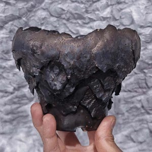 Puede incluir: Un cuenco de cerámica oscura y texturizada con una forma irregular y orgánica. El cuenco tiene un aspecto rugoso y en capas con un acabado oscuro y metálico. El cuenco se sostiene en una mano sobre un fondo gris texturizado. El cuenco parece hecho a mano.