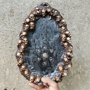 Puede incluir: Un plato de cerámica oscuro, de forma ovalada, con un borde de pequeñas calaveras detalladas. El interior tiene una superficie oscura y texturizada con varios agujeros pequeños. Las calaveras son de color marrón claro, contrastando con el interior oscuro.