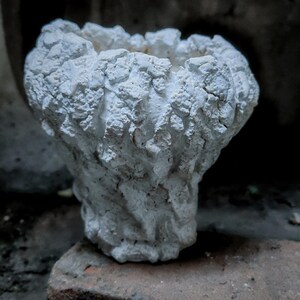 Puede incluir: Una maceta blanca texturizada, hecha a mano, con un aspecto rugoso y orgánico. La pequeña maceta decorativa tiene una amplia abertura y se apoya sobre una superficie de ladrillo. Ideal para plantas pequeñas o suculentas.