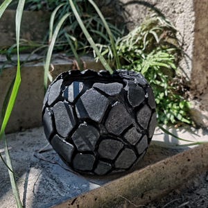 Può includere: Un vaso rotondo grigio scuro con una superficie strutturata a mosaico. Il vaso è realizzato in un materiale resistente ed è progettato per contenere piante. L'immagine mostra il vaso all'aperto, con piante verdi sullo sfondo.